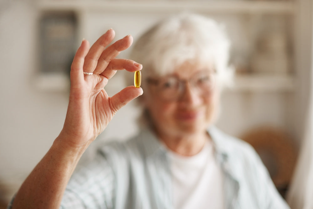 Close-up of an elderly woman’s hand holding a softgel capsule (fish oil / omega-3 supplement) over a table, preparing to take it with her meal