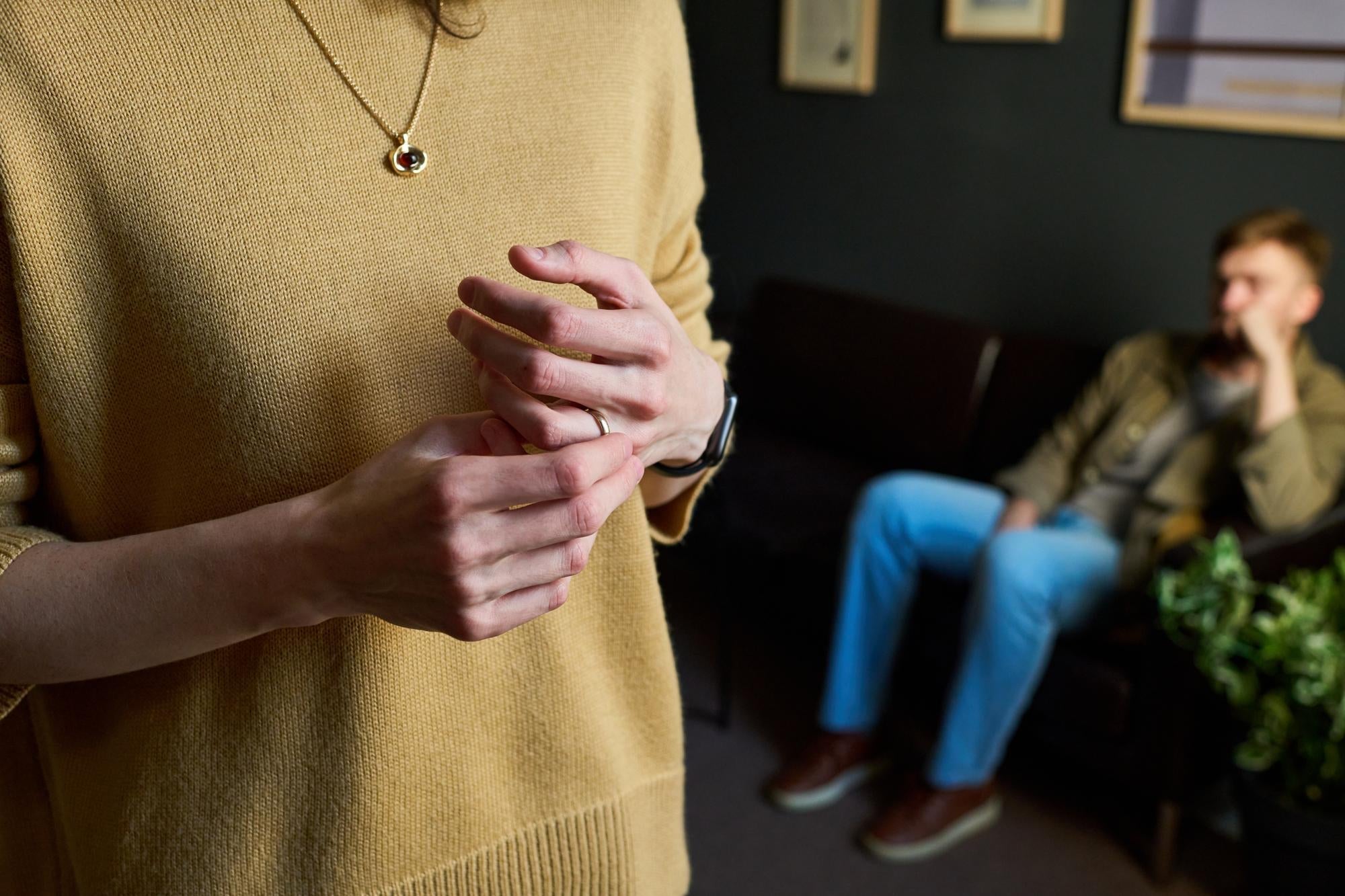 A person clasping their hands with visible tension, another individual seated apart in the background.