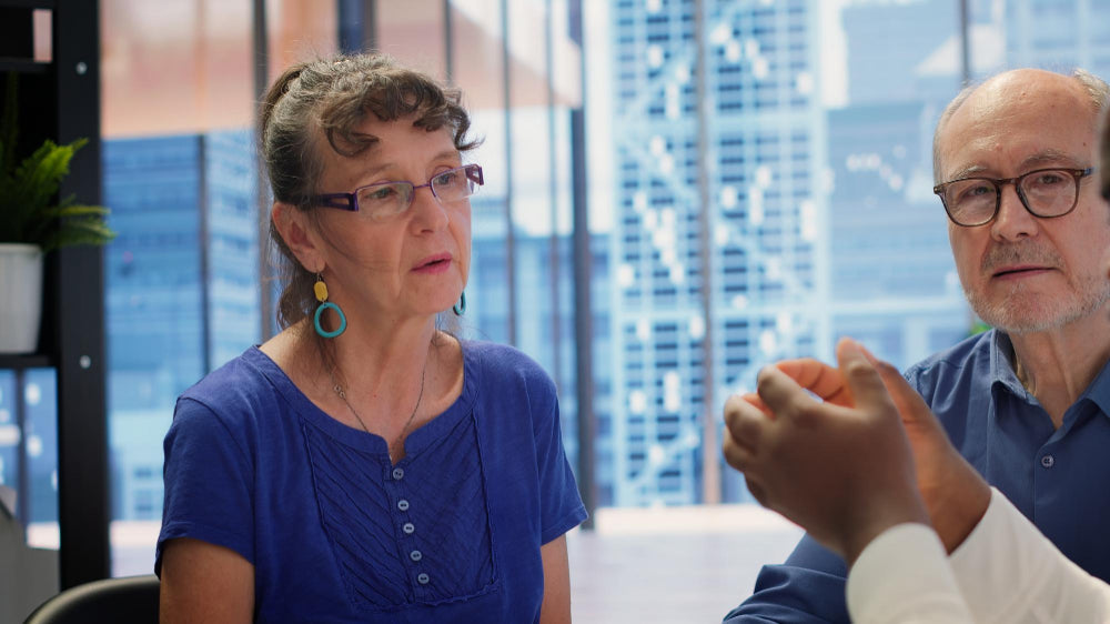 The senior couple is in a modern office and converses with a gesturing person.