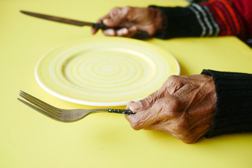 Senior woman holding cutlery next to an empty plate, illustrating the challenges faced by individuals with Parkinson’s disease during mealtime.