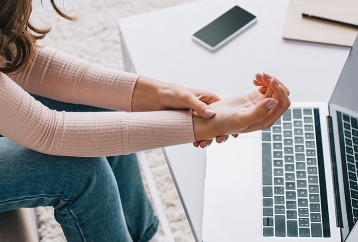 A person holding their wrist while sitting at a desk with a laptop, wondering why is my hand shaking and exploring the shaky hands causes.