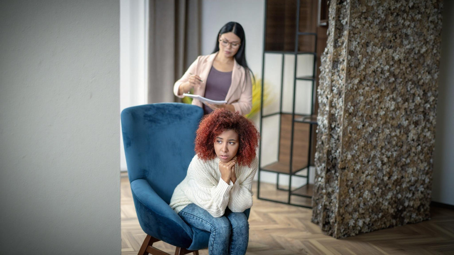 A young woman with curly hair sits across from her therapist, discussing stress-related symptoms such as anxiety tremors and psychogenic shaking. 