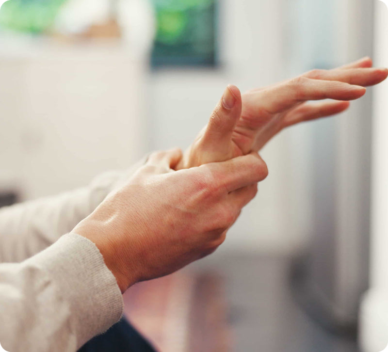 A close-up of a person's hands stretching or massaging their forearm, with a softly blurred indoor background.